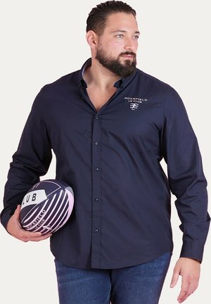 Chemise bleu marine à bouton en tissu tissé avec un ourlet arrondi, arborant le logo "RUCKFIELD LE CLUB". Le modèle tient un ballon de rugby rayé.