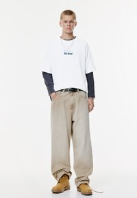 Ung person iført lagdelte hvide og sorte langærmede skjorte med teksten "Det er tid", beige løse jeans, brune støvler og sølvkæde tilbehør.