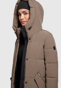 Hnedá prešívaná bunda s veľkou kapucňou, zapínaním na zips a patentkami. Má logo ako ozdobu a bočné vrecko pre funkčnosť.
