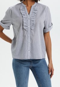 Blå och vit randig kortärmad skjorta med volangkant framtill och knäppning. Mjuk tygkvalitet, avspänt passform och uppvikta ärmar.
