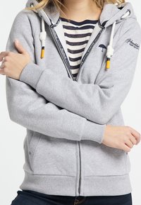 Felpa grigia con zip, dotata di lacci, polsini a costine e tasche laterali. Presenta un ombrello ricamato e una fodera interna a righe.