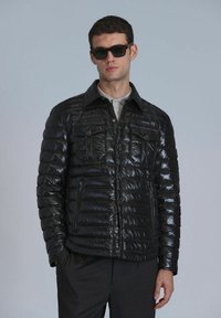 Schwarze Pufferjacke mit glänzender Oberfläche, zwei Brusttaschen vorne und Knopfverschluss. Das Modell trägt Sonnenbrillen und Hosen.