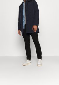 Marineblaue lange Jacke mit Kragen, mit Knopfleiste, getragen über einem hellblauen Hemd, schwarzen Hosen und weißen Sneakers mit blauen Akzenten.