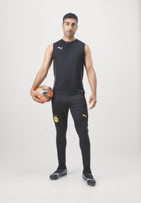 Zwarte mouwloze shirt en zwarte strakke broek met gele logo-accenten, houdend een oranje en zwarte voetbal, dragend voetbalschoenen.