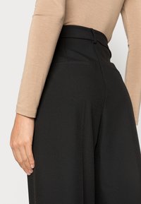 Kobieta ubrana w czarne, szerokie spodnie z wysokim stanem oraz beżową bluzkę z długim rękawem, z lewą ręką opartą wzdłuż ciała.