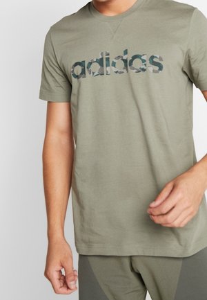 Mand iført olivengrøn Adidas T-shirt med mønstret logo over brystet, sat sammen med matchende grønne bukser.