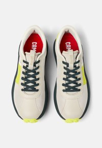 Sneakers beige con parte superiore in rete, lacci verdi, dettagli gialli brillanti e suola nera texturizzata. Soletta rossa con il logo del marchio visibile.