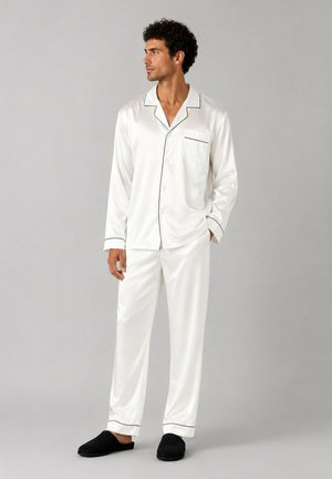 Homme portant un ensemble de pyjama en satin blanc avec des passepoils noirs et des pantoufles noires, debout devant un fond gris clair uni.