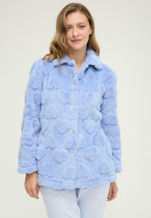 Joven mujer con una chaqueta azul claro y esponjosa con patrones de corazones en relieve y pantalones azul claro a juego con puntos blancos.