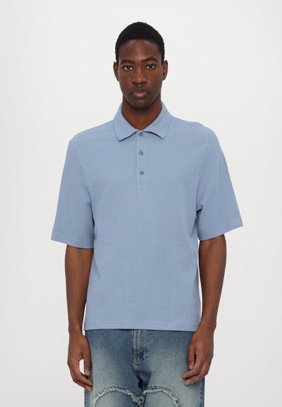 Un polo bleu clair à manches courtes avec un tissu texturé, un col classique et une patte de boutonnage à trois boutons, associé à un jean en denim déchiré.