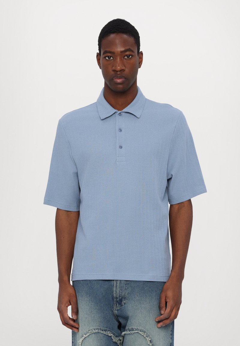 Šviesiai mėlyna trumpomis rankovėmis polo marškinėliai išsiskiria tekstūruotu audiniu, klasikine apykakle ir trimis mygtukais, derinami su nusidėvėjusiais džinsais.