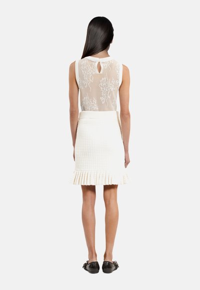 Haut en dentelle blanche avec design floral, détail en découpe au dos, et jupe blanche texturée avec ourlet à volants. Des ballerines noires complètent le look.
