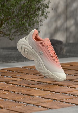 Baskets athlétiques rose clair et blanc avec une semelle épaisse flottant au-dessus d'un sol en bois et en carreaux de pierre à l'extérieur.