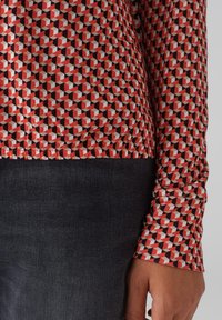 Langärmliges Shirt mit einem geometrischen Muster in Rot, Schwarz und Creme. Der Stoff ist glatt, mit einem leicht taillierten Schnitt und einem abgerundeten Saum.