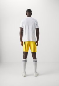 Camisa blanca de manga corta, pantalones cortos deportivos amarillos, calcetines blancos con rayas negras y botas de fútbol blancas, visto desde la parte trasera.