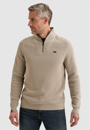 Hombre con suéter de punto beige acanalado con cremallera negra hasta un cuarto y vaqueros azul oscuro, mirando hacia un lado con un fondo liso.