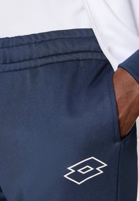 Marineblaue Sporthose mit glatter Textur, versehen mit weißen Nähten und einem weißen Logodetail am unteren linken Bein.