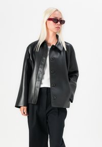 Black leather jacket with a button front, spread collar, and long sleeves. The model wears dark sunglasses and a white top.