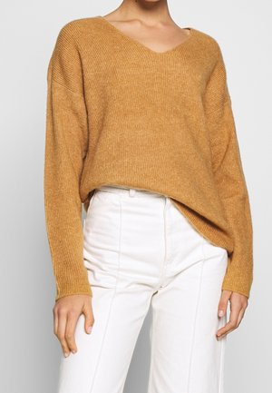 Personne portant un pull ample côtelé couleur camel avec un col en V et un pantalon blanc taille haute sur un fond gris.