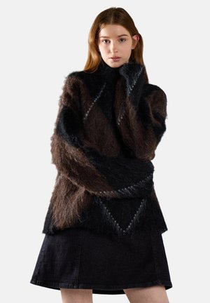 Jonge vrouw met lang haar, draagt een bruin-zwarte pluizige trui en een zwarte rok, poseert met één hand dicht bij haar gezicht.