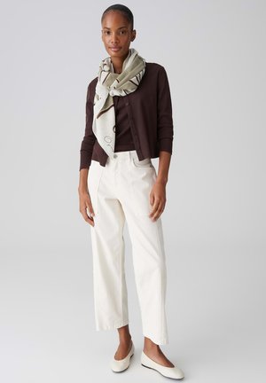 Frau trägt eine dunkelbraune Strickjacke, einen beigen Schal, cremeweiße Hosen mit weitem Bein und cremefarbene Ballerinas, steht vor einem hellgrauen Hintergrund.