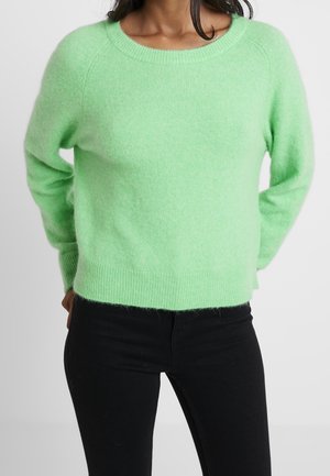 Femme portant un pull doux vert clair à manches longues et un pantalon noir, debout les mains dans le dos sur fond blanc.