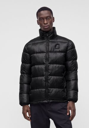 Homme portant une doudoune noire avec un col haut, fermeture éclair frontale, et un logo discret sur la poitrine gauche, debout devant un fond clair uni.