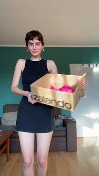Femeie într-o rochie neagră fără mâneci, ținând o cutie Zalando cu haine roz, stând într-un living cu pereți verzi și o canapea.