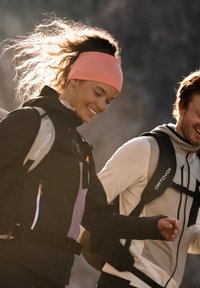 Two hikers wear technical outerwear. The woman has a pink headband and a black jacket with gray and purple accents. The man wears a light-colored jacket.