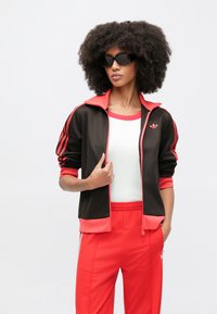 Czarna i czerwona kurtka dresowa na zamek z trzema paskami na rękawach, w zestawie z czerwonymi spodniami dresowymi i białym topem; logo akcentujące na piersi.