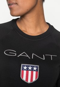 Sudadera negra con "GANT" bordado en gris sobre un emblema en forma de escudo con rayas rojas y blancas y detalles en azul. Cuello redondo.