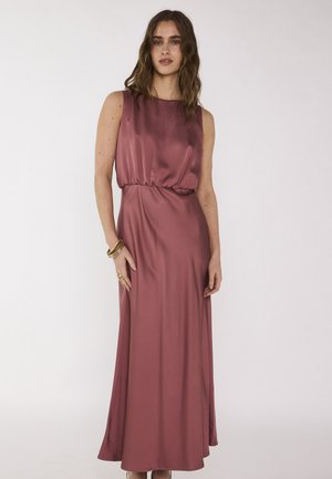 Vrouw draagt een mauve avondjurk van satijn zonder mouwen met een geplooide taille, staand tegen een effen lichte achtergrond, geaccentueerd met gouden armbanden.