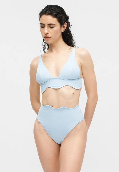 Femme aux cheveux foncés mouillés portant un bikini bleu clair à bord festonné, debout devant un fond gris clair uni, regardant vers le bas.
