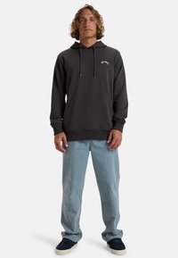 Dark gray hoodie with a front kangaroo pocket, adjustable drawstring hood, and subtle logo detail. Worn with light blue jeans and dark shoes.