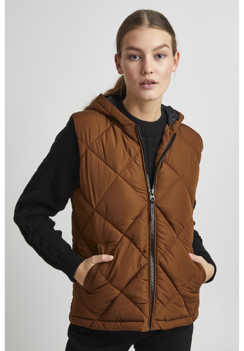Gilet matelassé marron avec capuche, comportant une fermeture éclair à l'avant et deux poches latérales. Les manches tricotées noires ajoutent de la texture et du contraste.