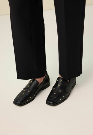 Mocassins en cuir noir ornés de clous dorés et d'abeilles, portés avec un pantalon noir raccourci sur un sol clair.