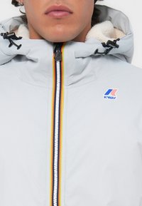 Lys grå vanntett jakke med glidelås og flerfargede striper. Fleece-fôret hette; K-Way-logo vist på brystet.