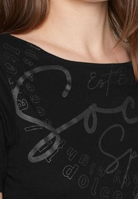 Černý krátký rukávový top s širokým výstřihem. Přední část zdobí lesklý černý text a abstraktní vzory různých velikostí a orientací.