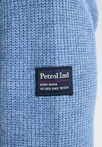 Blauwe gebreide stof met een gestructureerd patroon. Bevat een zwart etiket met witte en rode tekst, die merk- en modelinformatie aangeeft.
