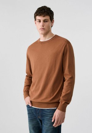Giovane uomo con capelli corti e ricci, che indossa un maglione marrone sopra una camicia bianca e jeans blu, in piedi davanti a uno sfondo grigio uniforme.