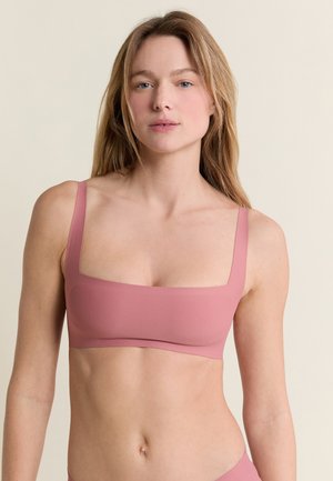 Femme aux longs cheveux châtain clair portant un soutien-gorge rose à encolure carrée sur un fond clair uni.