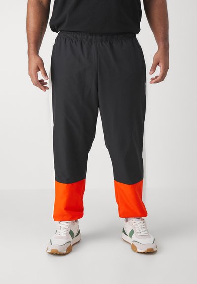Pantalons de sport noirs avec un large panneau inférieur orange, une taille élastique et des bandes latérales. Portés avec des baskets blanches et vertes.