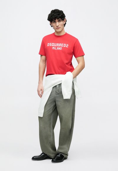 Punane t-särk valge kirjaga "DSQUARED2 MILANO", kombineeritud lahtiste roheliste teksade ja mustade kingadega. Vöökoha ümber on seotud valge kampsun.