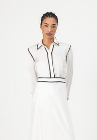 Robe blanche avec des bordures noires, manches longues, fermeture à boutons à l'avant et taille cintrée. Tissu lisse avec une jupe fluide.