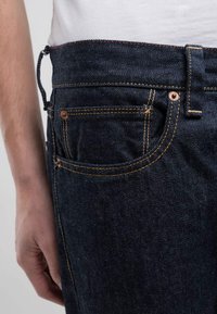 Jeans de mezclilla oscura con costuras naranjas contrastantes, diseño de cinco bolsillos y herrajes de cobre. Incluye una línea decorativa roja en la cinturilla.