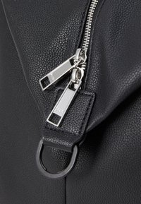 Sac en cuir noir texturé avec des doubles zips argentés, des tirettes rectangulaires et un détail en anneau métallique attaché à la bandoulière.