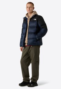 Blue and black puffer jacket with a zippered front, worn over a beige hoodie and paired with olive green pants and black sneakers.