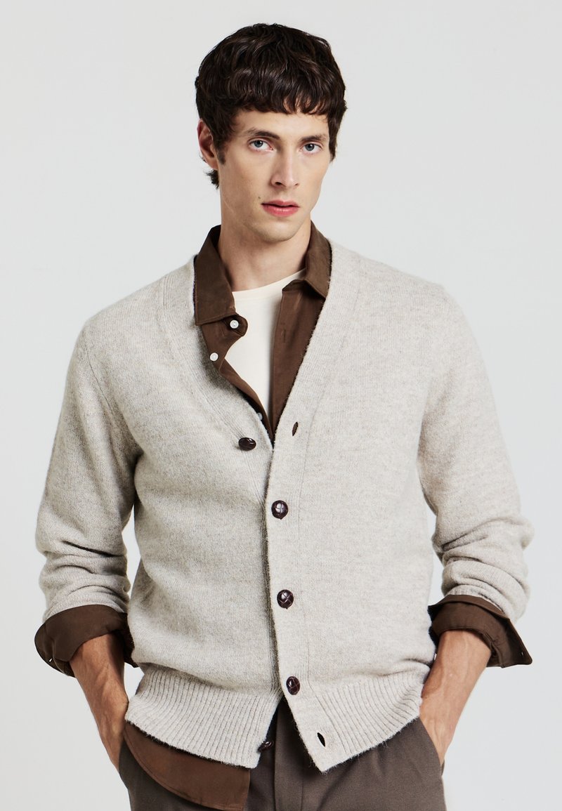 Beige Strickjacke mit V-Ausschnitt, ausgestattet mit dunklen Knöpfen und gerippten Bündchen. Über einem braunen Hemd mit Knöpfen getragen.