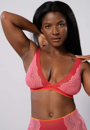 Roze kant bralette met doorschijnende stof, driehoekige cups en gele onderband. Model toont het ontwerp met één arm achter haar hoofd.