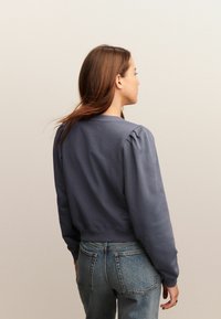 Vrouw met lang bruin haar, gekleed in een grijze top met pofmouwen en een blauwe spijkerbroek, staande met haar rug naar een effen beige achtergrond.
